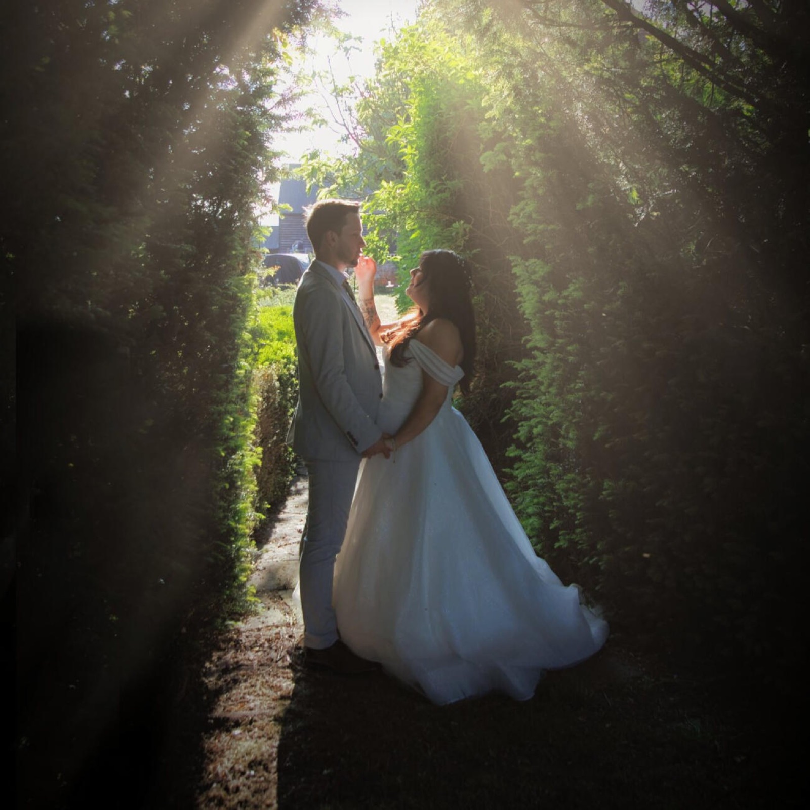 Bride and groom facing each other in a hedge-lined garden path, lit by rays of sunlight